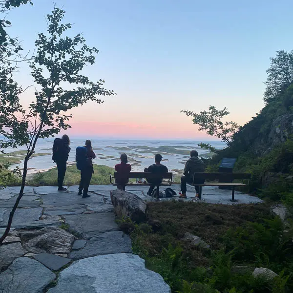 Scenic coastal lookout featuring visitors, seating areas and panoramic views of an archipelago at sunset, integrated into a natural landscape setting.