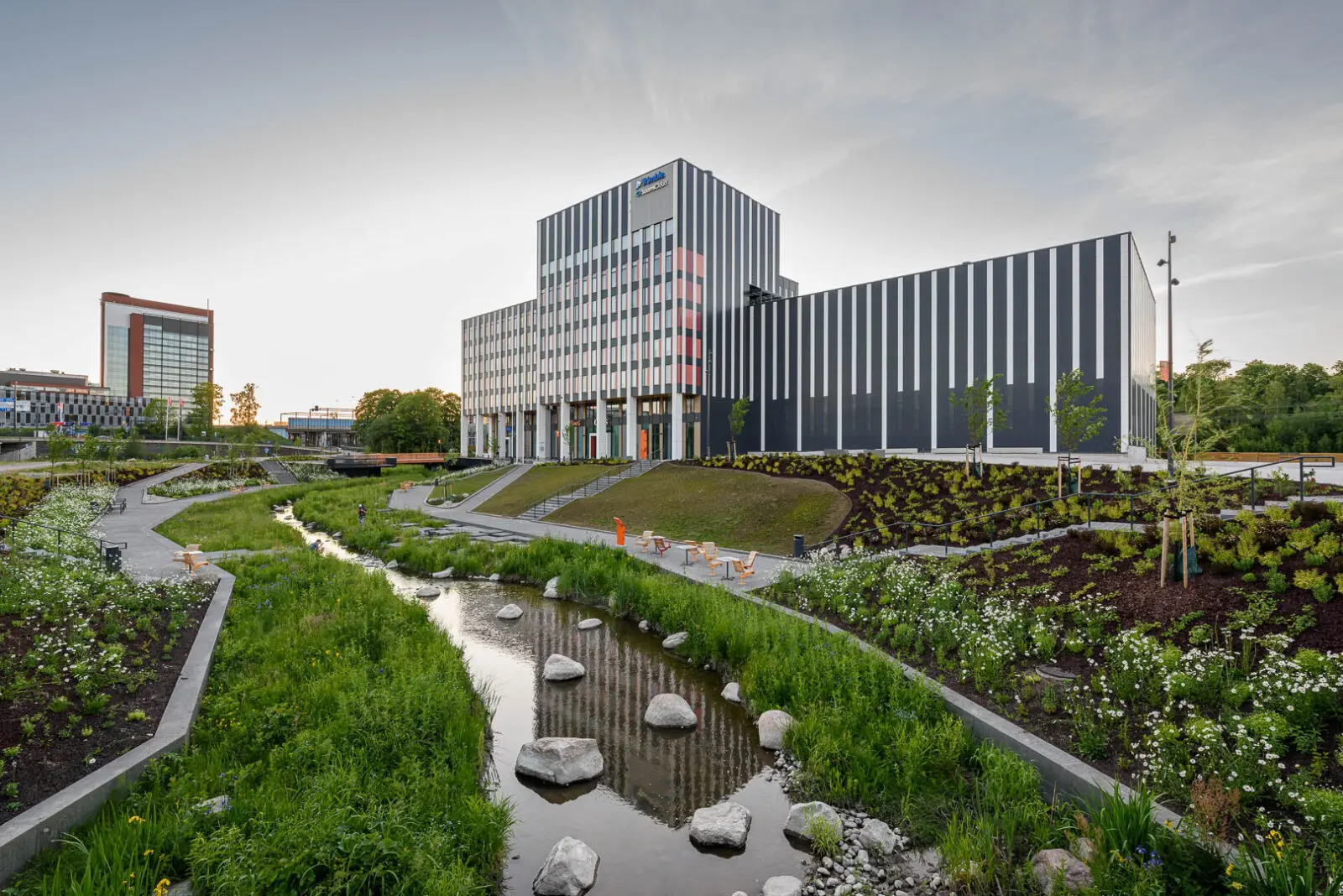 Blue-green urban landscape combining a naturalized watercourse, biodiversity-rich planting, sustainable drainage systems, and accessible public spaces within a contemporary business district.
